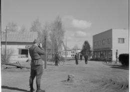 Kuvassa etualalla valokuvaa ottava sotilas. Kuvassa myös ihmisiä ja liikerakennuksia.