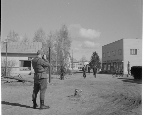 Kuvassa etualalla valokuvaa ottava sotilas. Kuvassa myös ihmisiä ja liikerakennuksia.