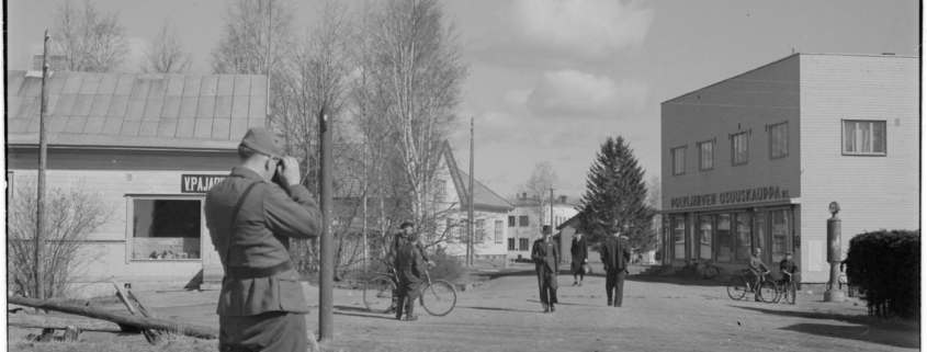 Kuvassa etualalla valokuvaa ottava sotilas. Kuvassa myös ihmisiä ja liikerakennuksia.