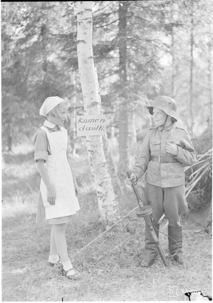 Kuvassa pikkulotta ja sotilaspoika hymyilevät toisilleen. Sotilaspojalla on kädessä Suomi-konepistooli.