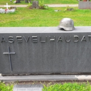 Kuvassa hautakivi, jossa teksti asevelihaudat. Kivessä myös risti ja kiven päällä sotilaan kypärä.