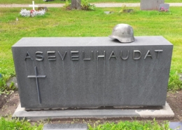 Kuvassa hautakivi, jossa teksti asevelihaudat. Kivessä myös risti ja kiven päällä sotilaan kypärä.