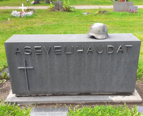 Kuvassa hautakivi, jossa teksti asevelihaudat. Kivessä myös risti ja kiven päällä sotilaan kypärä.