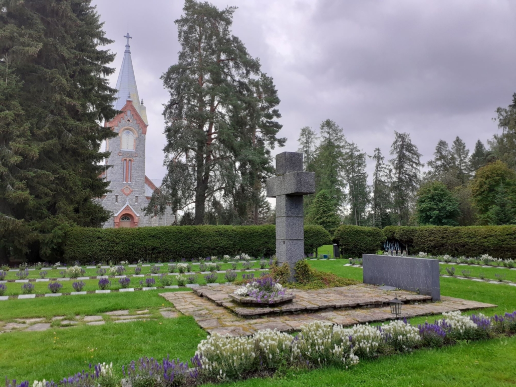 Värikuvassa sankarihautausmaan suuri kiviristi ja hautoja sekä kirkon torni.