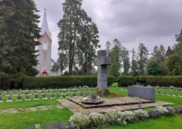 Värikuvassa sankarihautausmaan suuri kiviristi ja hautoja sekä kirkon torni.