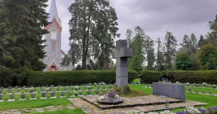 Värikuvassa sankarihautausmaan suuri kiviristi ja hautoja sekä kirkon torni.