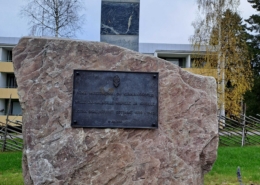 Kuvassa suurikokoinen muistomerkkikivi, jossa tekstilaatta ja Suomen sinivalkoinen rajapaalu.