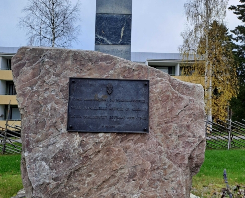 Kuvassa suurikokoinen muistomerkkikivi, jossa tekstilaatta ja Suomen sinivalkoinen rajapaalu.