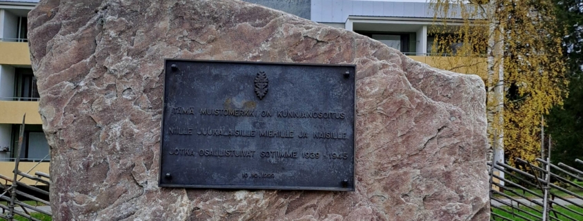Kuvassa suurikokoinen muistomerkkikivi, jossa tekstilaatta ja Suomen sinivalkoinen rajapaalu.