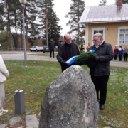 Kuvassa kaksi miestä laskemassa seppelettä muistomerkille. Yleisönä mm. oppilaita.