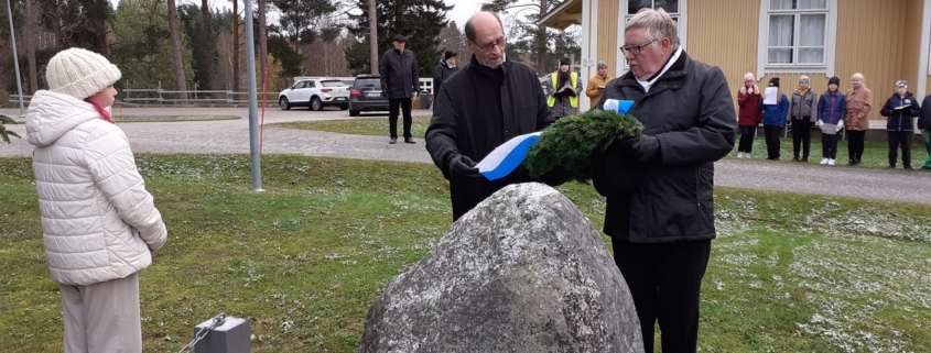 Kuvassa kaksi miestä laskemassa seppelettä muistomerkille. Yleisönä mm. oppilaita.