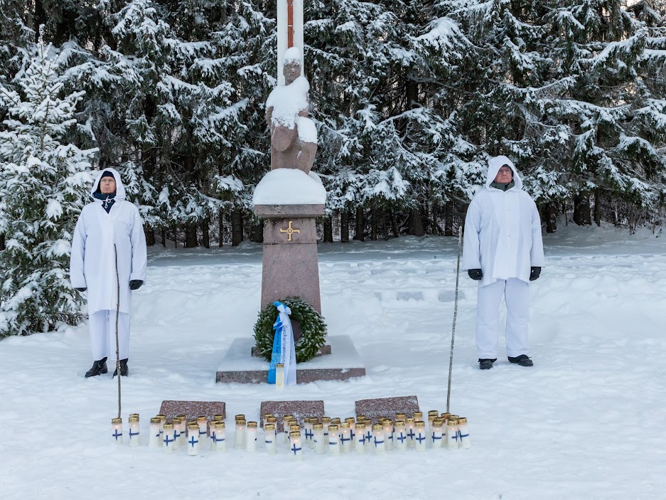 Kuvassa lumipukuiset sotilaat seisovat kunniamerkin vierellä. Edessä seppele ja kynttilöitä.