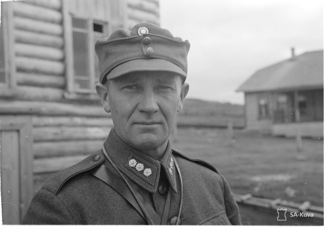 Mustavalkoisessa kuvassa everstinarvoinen sotilaspukuinen mies. Taustalla hirsirakennuksia