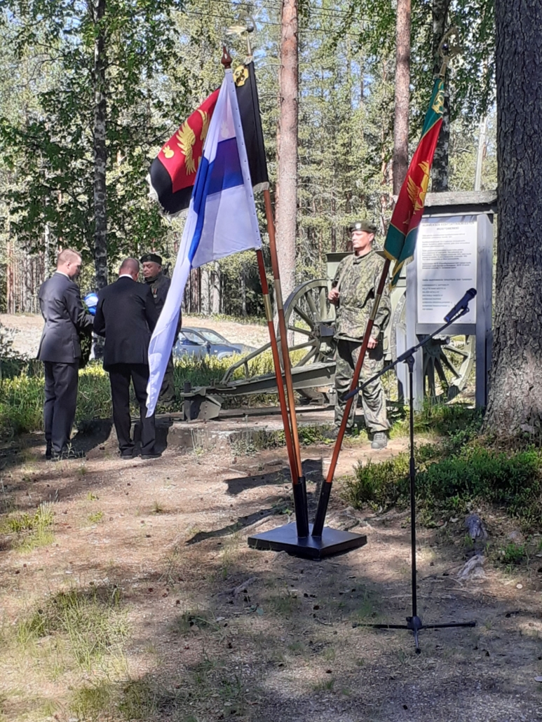 Kaksi miestä laskemassa seppelettä muistomerkille, jossa kunniavartio. Etualalla lippuja.