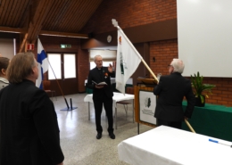 Kuvassa mies mustassa puvussa kantaa perinneyhdistyksen lippua ja toinen mies siunaa lippua. Lippu on valkoinen ja siinä on vihreä tammenlehvä-kuvio.