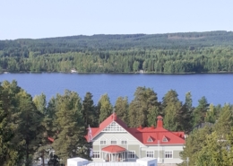 Kuvassa kansallisromanttinen puurakennus, jonka taustalla järvi ja vaaroja.