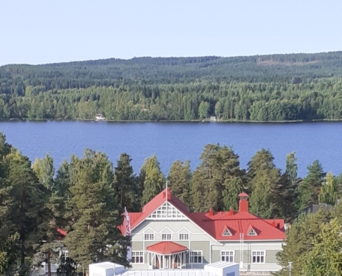 Kuvassa kansallisromanttinen puurakennus, jonka taustalla järvi ja vaaroja.