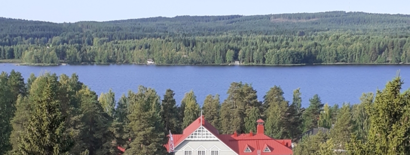 Kuvassa kansallisromanttinen puurakennus, jonka taustalla järvi ja vaaroja.