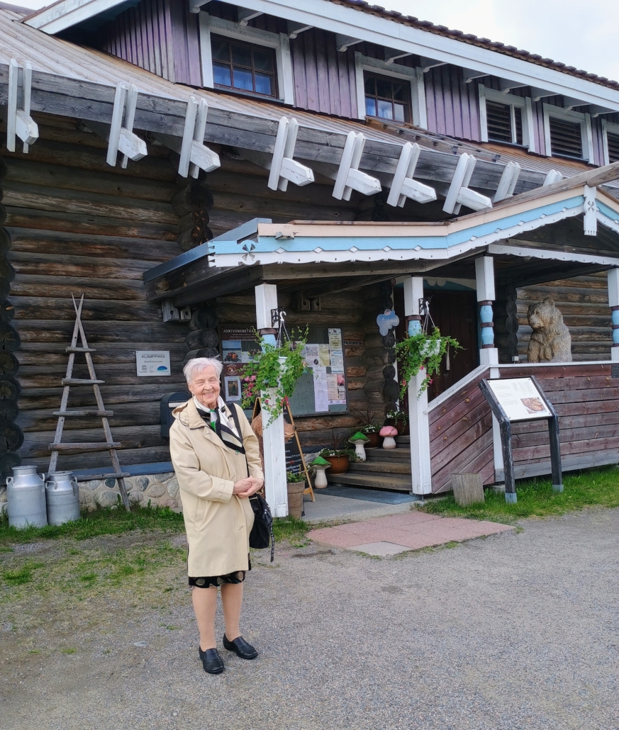 Kuvassa nainen ja taustalla karjalaistyylinen rakennus.