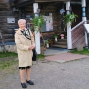 Kuvassa nainen ja taustalla karjalaistyylinen rakennus.