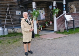 Kuvassa nainen ja taustalla karjalaistyylinen rakennus.