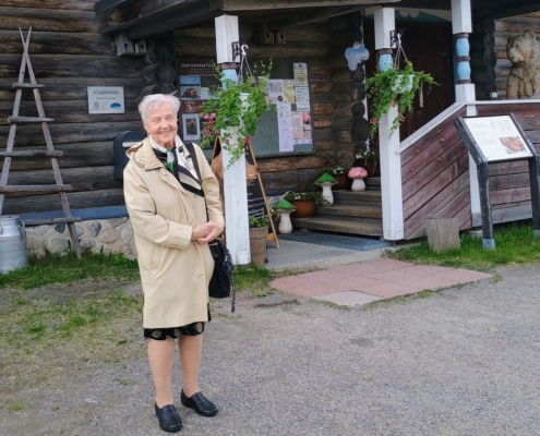 Kuvassa nainen ja taustalla karjalaistyylinen rakennus.