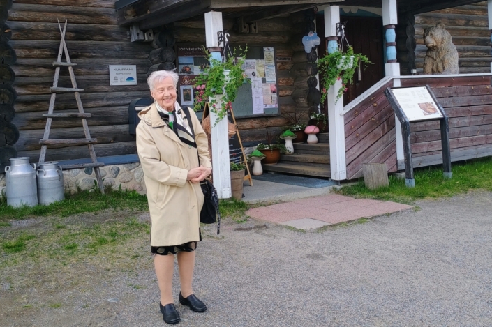 Kuvassa nainen ja taustalla karjalaistyylinen rakennus.