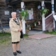 Kuvassa nainen ja taustalla karjalaistyylinen rakennus.