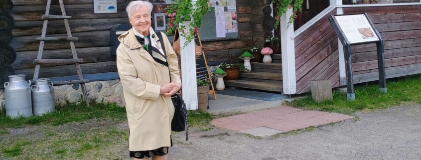 Kuvassa nainen ja taustalla karjalaistyylinen rakennus.