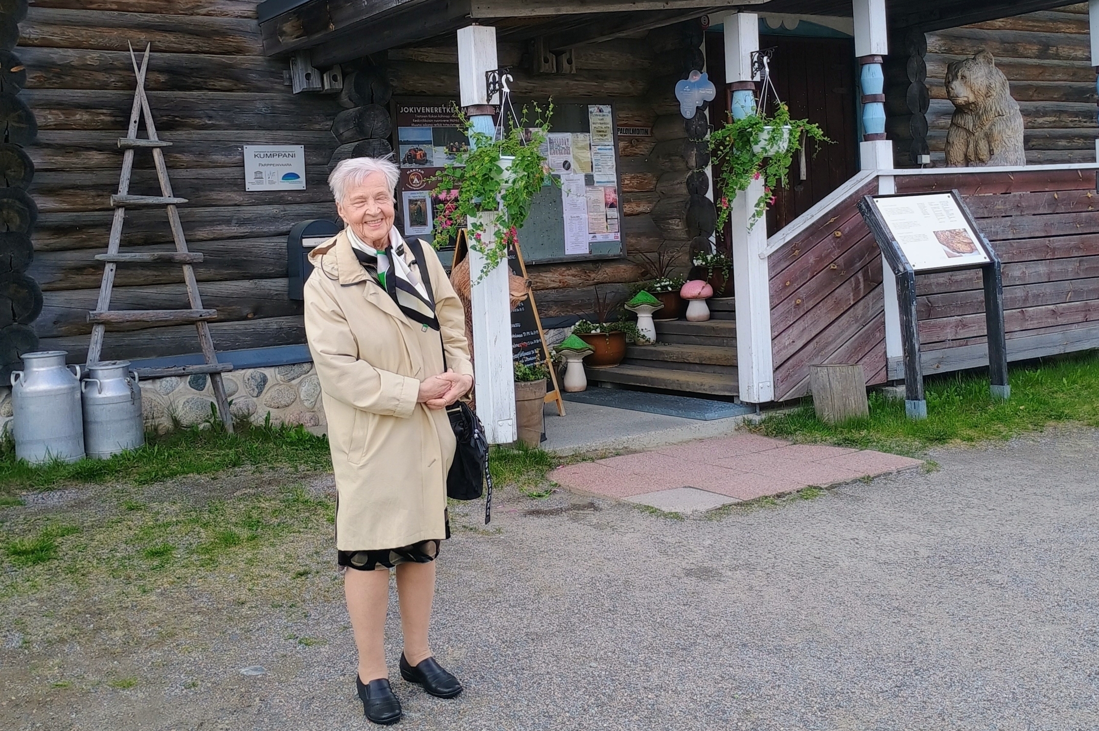 Kuvassa nainen ja taustalla karjalaistyylinen rakennus.