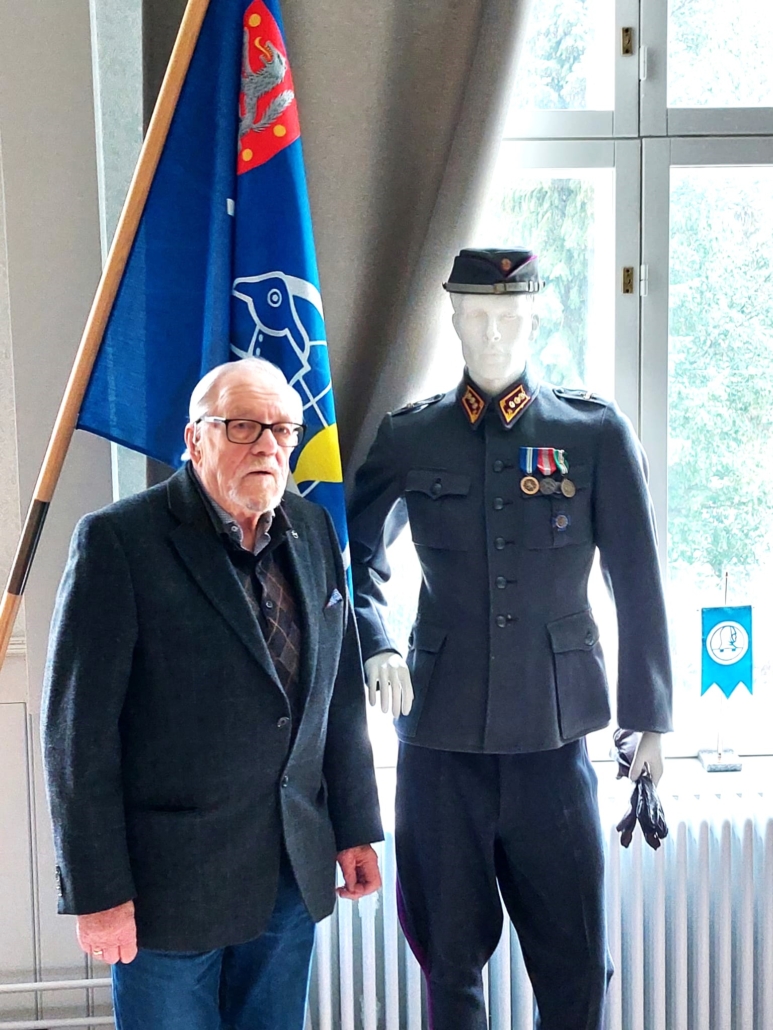 kuvassa mieshenkilö sotilaspuvun vieressä. Taustalla veteraanijärjestön lippu.