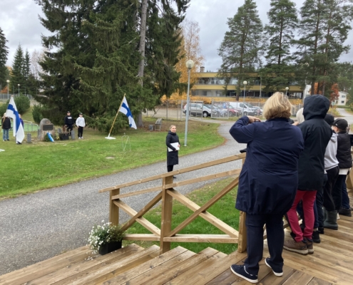 Kuvassa tilaisuuden osallistujia, muistomerkki ja Suomen liput.