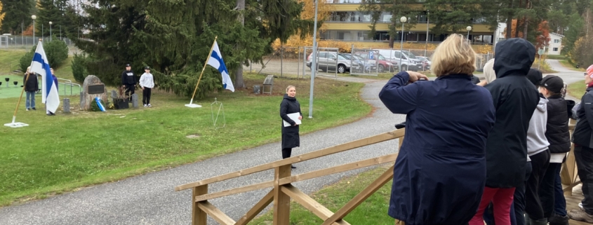 Kuvassa tilaisuuden osallistujia, muistomerkki ja Suomen liput.