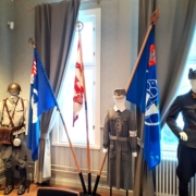 Kuvassa sotilaspukuja ja veteraanijärjestöjen lippuja jalustassa.