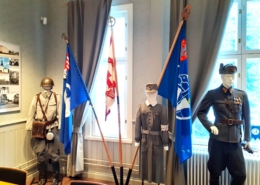 Kuvassa sotilaspukuja ja veteraanijärjestöjen lippuja jalustassa.