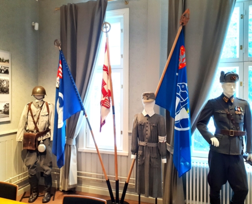 Kuvassa sotilaspukuja ja veteraanijärjestöjen lippuja jalustassa.