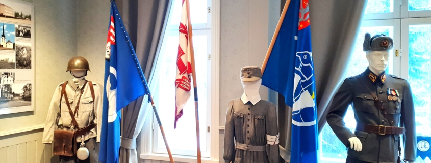 Kuvassa sotilaspukuja ja veteraanijärjestöjen lippuja jalustassa.