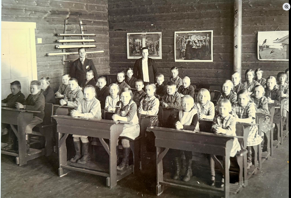Mustavalkoisessa kuvassa oppilaita kansakoululuokassa pulpettien ääressä. Taustalla seisoo kaksi opettajaa.