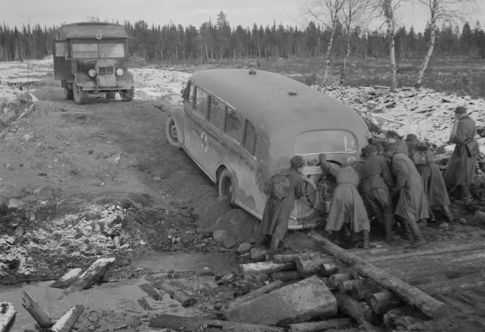 Sairaskuljetusbussi autetaan hinaamalla ja miesvoimin työntämällä rikotulla Jäämeren tiellä.